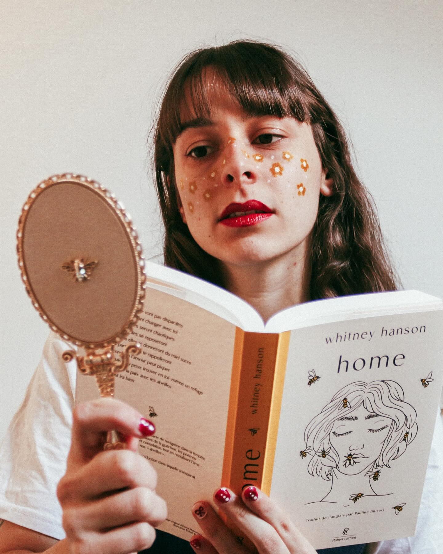 Solenne, fleurs dorées peintes sur les joues, lit le recueil « Home » de Whitney Hanson en tenant un miroir à main.